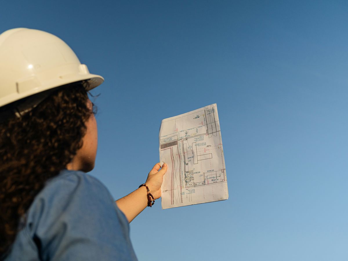 Een vrouw met een helm houdt een bouwplan omhoog tegen de blauwe lucht